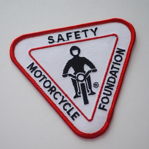 May include: Embroidered patch in a triangular shape with a red border. The patch features the words "SAFETY," "MOTORCYCLE," and "FOUNDATION." A black silhouette of a motorcyclist on a motorcycle is in the center.