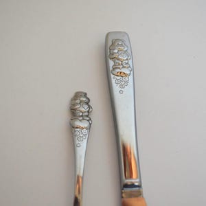 May include: Two silver-toned metal utensils featuring embossed Care Bear character designs on the handles. The smaller utensil on the left and the larger utensil on the right both display a bear figure standing on a cloud with small flower accents.