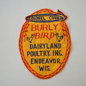 Może przedstawiać: Winylowa żółta naszywka z czerwonym i niebieskim haftowanym tekstem i grafiką. Na naszywce widnieje napis "COLONEL CORN" u góry, "BURLY BIRD" na środku i "DAIRYLAND POULTRY, INC. ENDEAVOR, WIS." na dole. Po obu stronach tekstu znajdują się dwa niebieskie kurczaki.