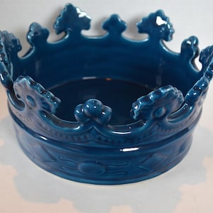 以下が含まれることがあります： 濃いティール色のセラミック製の王冠型ボウル。ボウルは幅が広く浅い内側と、縁に沿った詳細な王冠のデザインが特徴です。王冠は装飾的なディテールと光沢のある仕上げが施されています。