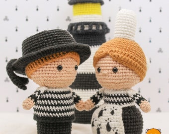 May include: Two crocheted amigurumi dolls, one wearing a black and white striped shirt and a black hat, the other wearing a white and black dress with a brown hairpiece. The dolls are standing in front of a black and white striped lighthouse.