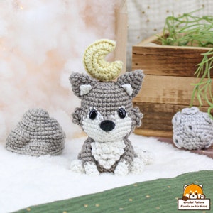 Puede incluir: Un juguete de amigurumi de lobo gris tejido a crochet con una luna creciente amarilla en la cabeza. El lobo tiene pelo blanco en el pecho y una nariz negra. El juguete está sentado sobre una superficie blanca y esponjosa.