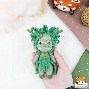 May include: A green crocheted amigurumi doll with a leafy head and a green dress. The doll has black eyes and a small flower on its head.