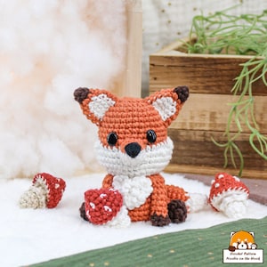 Puede incluir: Un zorro de crochet naranja con detalles blancos se sienta sobre una superficie blanca con tres setas de crochet rojas y blancas. El zorro tiene ojos negros y una nariz negra.