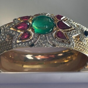 Op de afbeelding: Een goudkleurige scharnierende armband met een centrale smaragdgroene edelsteen, omringd door rode en heldere edelstenen. De armband heeft een getextureerde band en ligt in een witte doos.