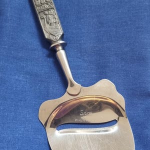 Puede incluir: Un cortador de queso de acero inoxidable con un mango decorativo. El mango presenta tallas en relieve detalladas de figuras y símbolos. El cortador tiene una hoja curva y la palabra "Spor" está grabada en la hoja.