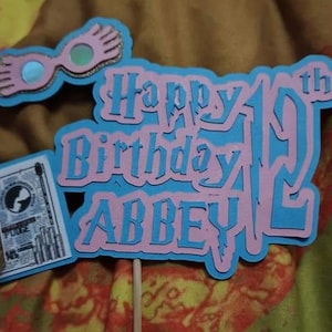 May include: A pink and blue cake topper with the text "Happy 19th Birthday ABBEY". The topper features a pair of glasses with wings, a book, and a stick for inserting into a cake.
