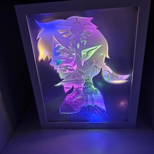May include: A white framed light-up art piece featuring a silhouette of a person with a landscape behind them. The silhouette is lit with a blue, green, and purple light. The landscape is lit with a pink and purple light. The person is holding a sword and has a triangular symbol on their head.