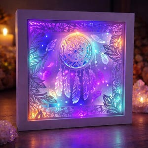 May include: A white framed dreamcatcher art piece with a layered design. The artwork features a dreamcatcher in the centre, surrounded by feathers and leaf patterns, illuminated by colourful LED lights in shades of purple, blue, and orange.