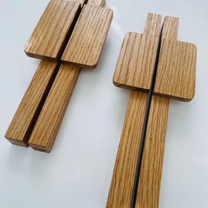 Op de afbeelding: Twee lichtbruine houten telefoonstandaards. Elke standaard heeft een rechthoekige bovenkant met een verticale sleuf en twee verticale steunen. De standaards zijn gemaakt van hout en hebben een natuurlijke houtnerf. Ze zijn ongeveer 15 cm lang.