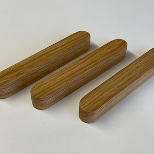 Peut inclure: Trois blocs de bois rectangulaires aux extrémités arrondies. Les blocs sont en bois brun clair et sont disposés horizontalement sur une surface blanche. Le grain du bois est visible sur la surface des blocs.