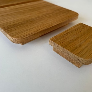 Puede incluir: Primer plano de dos piezas de madera sin terminar, de color marrón claro. Una pieza es rectangular y la otra es un cuadrado más pequeño. La veta de la madera es visible y los bordes son redondeados. Las piezas parecen ser parte de una estructura de madera más grande.
