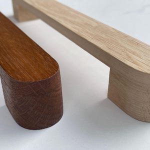 Pode incluir: Duas puxadores de armário de madeira, uma é de madeira marrom claro e a outra é de madeira marrom escuro. Ambas as puxadores têm uma forma retangular arredondada.