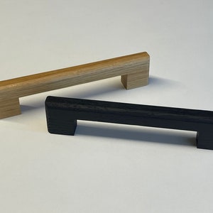 Op de afbeelding: Twee houten kastgrepen, een van naturel hout en een van zwart gebeitst hout. Beide grepen hebben een rechthoekige vorm met een uitgesneden handvat.