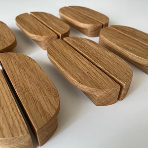 Può includere: Diversi supporti per telefono in legno sono disposti su una superficie bianca. Ogni supporto, in legno chiaro, presenta una fessura per sostenere un telefono. I supporti hanno una forma arrotondata e venature naturali.