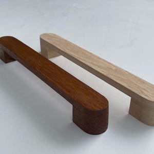 Può includere: Due maniglie per armadi in legno, una in legno marrone chiaro e l'altra in legno marrone scuro. Entrambe le maniglie hanno una forma rettangolare arrotondata.