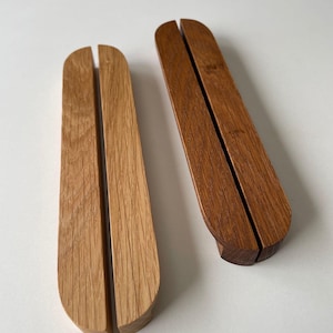 May include: Two wooden phone stands. One is light-colored, the other is dark. Each stand has a curved top and a slot down the center to hold a phone. The stands are made of wood and have a smooth finish.