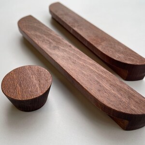 Könnte beinhalten: Drei Holzmöbelbeschläge: zwei lange, gebogene Griffe und ein runder Knopf. Die Teile sind aus gebeiztem Holz in einem satten, dunkelbraunen Farbton gefertigt. Die Griffe sind etwa 30 cm lang, und der Knopf hat einen Durchmesser von etwa 4 cm.