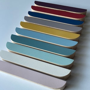 Puede incluir: Una colección de piezas de madera, cada una con un borde curvo y pintada en un color liso diferente. Los colores incluyen tonos de morado, rojo, azul, gris, amarillo y varios azules y verdes. Las piezas están dispuestas en diagonal.