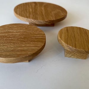 Pode incluir: Três puxadores de armário de madeira de carvalho com acabamento natural. Os puxadores têm formato oval e arredondado com base plana. O veio da madeira é visível, adicionando textura. Adequados para gavetas ou armários.