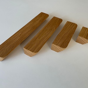 Könnte beinhalten: Vier Holzschrankgriffe mit natürlicher Eichenholzoberfläche. Die Griffe sind rechteckig mit abgewinkelten Enden, in verschiedenen Größen. Die Holzmaserung ist sichtbar. Die Griffe sind diagonal auf einer weißen Oberfläche angeordnet.
