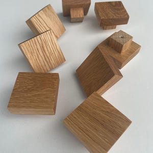 Może przedstawiać: Osiem kwadratowych drewnianych gałek do szafek w jasnobrązowym wykończeniu. Gałki mają kwadratowy kształt i są wykonane z litego drewna.