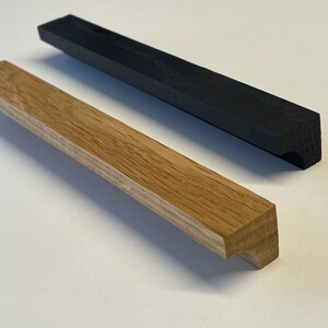Può includere: Due maniglie per ante di armadio in legno, una in legno naturale e una in legno tinto di nero. Entrambe hanno una forma rettangolare con un leggero angolo all'estremità.