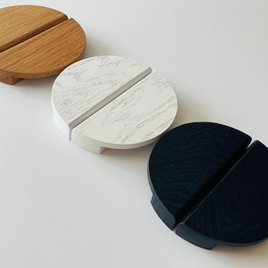 Peut inclure: Trois supports de téléphone en bois. Le premier est brun clair, le second est blanc avec des marbrures grises et le troisième est noir. Chacun a une fente pour un téléphone et une petite base.