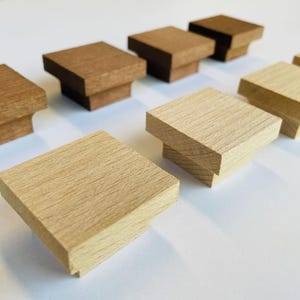 May include: A collection of square wooden cabinet knobs in various shades of brown and natural wood tones. The knobs have a stepped design, with a larger square top and a smaller square base. The knobs are arranged on a white surface.
