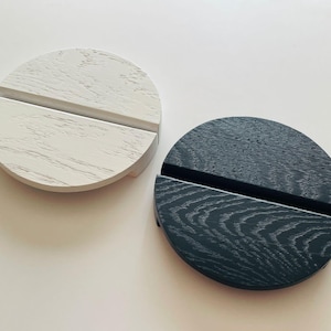 Op de afbeelding: Twee houten telefoonstandaards, één wit en één donkergrijs, elk met een sleuf voor het vasthouden van een telefoon. De standaards zijn rond met een platte bodem en een getextureerde houtnerf afwerking. De standaards staan op een witte ondergrond.