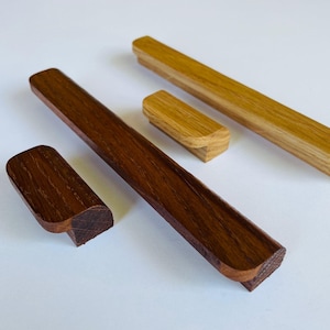 Può includere: Un assortimento di maniglie e pomelli per mobili in legno di varie forme e dimensioni. Le maniglie sono rettangolari e i pomelli arrotondati, tutti con finitura a venatura naturale. Le tonalità del legno variano dal rovere chiaro al noce scuro.