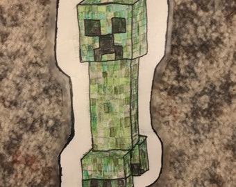 May include: A hand-drawn sticker featuring a Minecraft Creeper character. The Creeper is primarily green with a pixelated design, black facial features, and a rectangular body. The sticker has a white border and a signature in the bottom left corner.