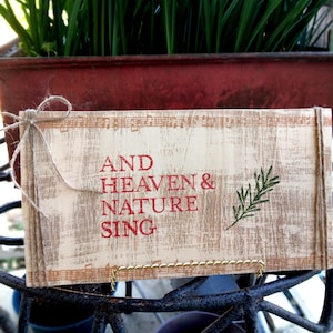 Puede incluir: Un letrero rectangular de madera con las palabras "AND HEAVEN & NATURE SING" en letras rojas. El letrero tiene un aspecto envejecido y rústico con una ramita de hojas verdes. Está atado con hilo y se exhibe en un soporte de metal.