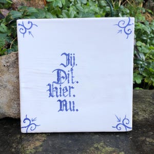 Op de afbeelding: Vierkante witte keramische tegel met blauwe letters en hoekontwerpen. De tekst luidt "Jij. Dit. Hier. Nu." in een decoratief lettertype. De tegel is waarschijnlijk voor decoratieve doeleinden.
