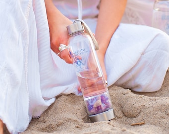 May include: A clear glass water bottle with a silver lid and a silver base. The bottle is filled with water and purple crystals. The bottle has a label that says "Gemstone Energy Bottle".