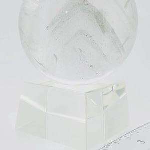 May include: A clear, spherical crystal ball resting on a faceted, transparent square base. A white ruler is positioned in the foreground, showing the base width is approximately 2 inches and the total height is over 3 inches.