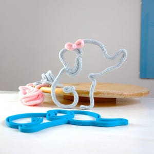 Puede incluir: Un elefante de punto gris con un lazo rosa en la cabeza. El elefante está sobre una base de madera. Hay una bola de hilo rosa y una forma de elefante de plástico azul sobre la mesa.