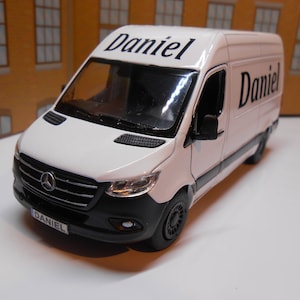 May include: White Mercedes-Benz Sprinter van toy model with black detailing. The van has the name "Daniel" printed on the side and the front of the van.