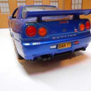 May include: A blue scale model Nissan Skyline car featuring a rear spoiler, circular red taillights, and a yellow number plate with the text DAN 17. The vehicle has dual exhaust pipes and the word SKYLINE embossed on the boot lid in silver lettering.