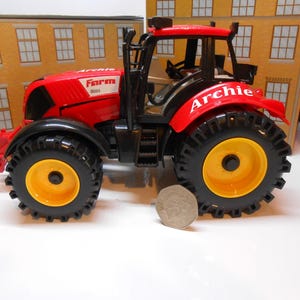 May include: A red toy tractor with black tyres and yellow wheel centres. The tractor has the name "Archie" on the side and "Farm 3000" on the front. A coin is next to the tractor.