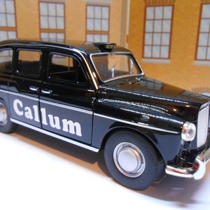 May include: A black toy London taxi with the name "Callum" in white lettering on the side. The miniature vehicle has silver accents, a taxi sign on top, and is set against a backdrop of a building.