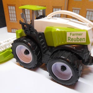 May include: Green and white toy tractor with a black and white rolling cutter. The tractor has a white sign that says "Farmer Reuben".