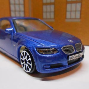 May include: A blue toy car with a metallic finish. The car has black and white wheels and a front license plate that reads "MICHAEL". The car is a coupe style.