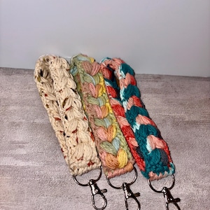 Puede incluir: Tres llaveros de muñeca tejidos a ganchillo con cierres de mosquetón de metal plateado. El primero es crema con motas multicolores, el segundo presenta un patrón trenzado en rosa pastel, amarillo y azul, y el tercero es una mezcla trenzada de verde azulado, rojo y rosa.