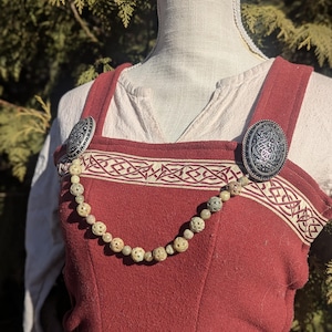 Puede incluir: Un vestido rojo de estilo medieval con adornos color crema y broches plateados decorativos. Un collar de cuentas con cuentas esféricas talladas cuelga sobre el pecho. El vestido se exhibe en un maniquí.