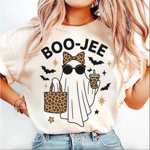 May include: A cream-coloured t-shirt with a Halloween graphic. The design shows a ghost wearing sunglasses and a bow, holding a leopard print tote bag and a coffee cup. The text "BOO-JEE" is above the ghost, with bats and stars.
