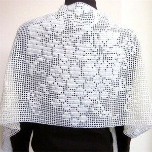 May include: A white, open-weave crochet shawl. The shawl is draped over a person's shoulders, showcasing a detailed floral pattern in the center. The edges have a delicate, lacy appearance. The shawl is made of a soft, lightweight material.