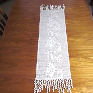 May include: White crocheted table runner with a floral design and fringe.