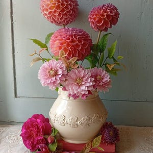 May include: A cream-colored ceramic vase with a relief leaf pattern around the center, filled with a bouquet of pink and coral dahlia flowers. Additional dahlia blossoms and green leaves are arranged around the base of the vase on a lace cloth.