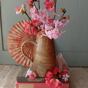 Peut inclure: Un pichet en céramique marron rempli de fleurs roses et rouges et un éventail décoratif. Le pichet est posé sur une pile de livres anciens aux couvertures roses et vertes. Un tissu rose clair est drapé à proximité.